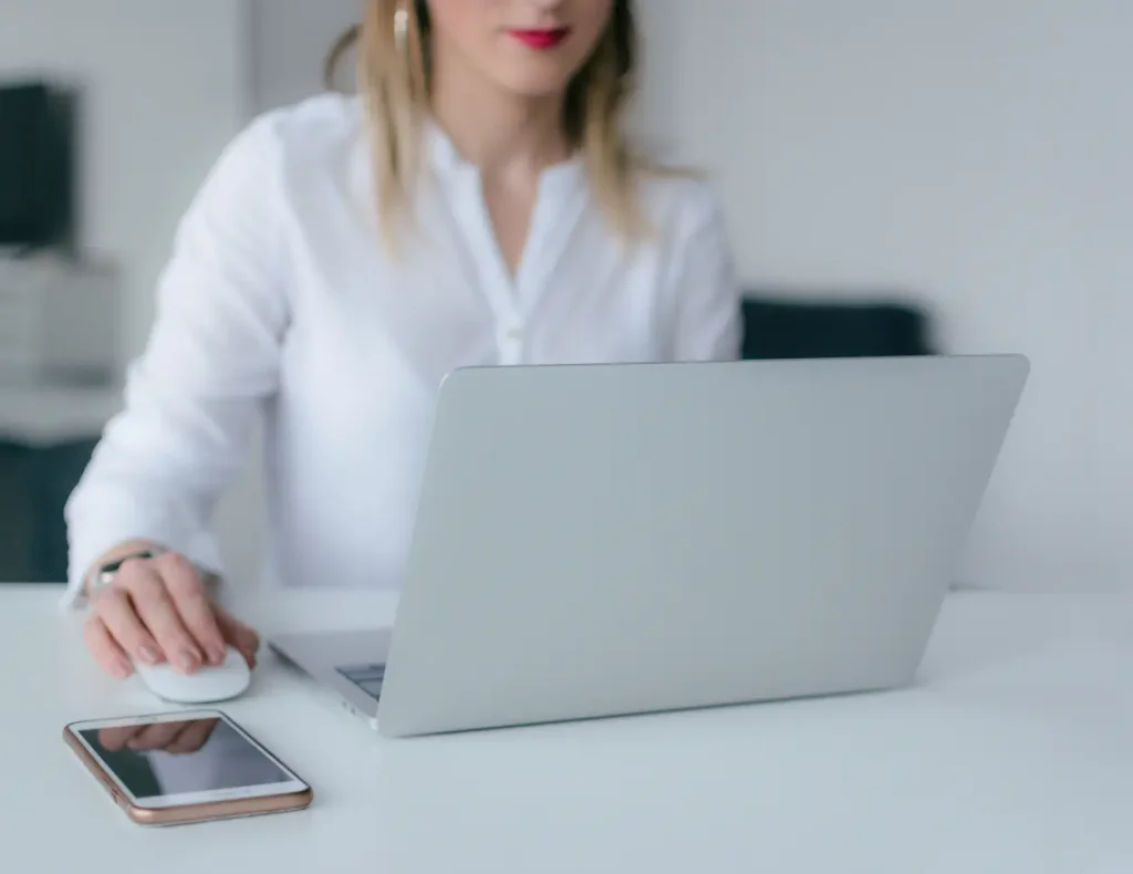 Woman working on laptop with smartphone next to her on a white desk for the service of Digital Agency Jinni S.r.l.