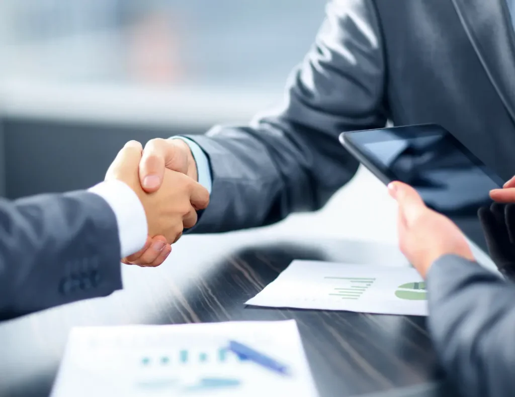 Two men in jackets shake hands at a table with documents and tablets.